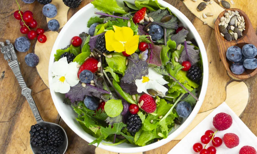 Bunte Salatschüssel mit Beeren und essbaren Blüten auf einem Holztisch.