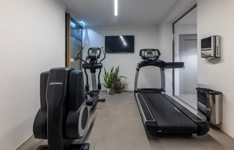 Fitnessbereich mit zwei Laufgeräten und einem Bildschirm, Pflanzen im Hintergrund
