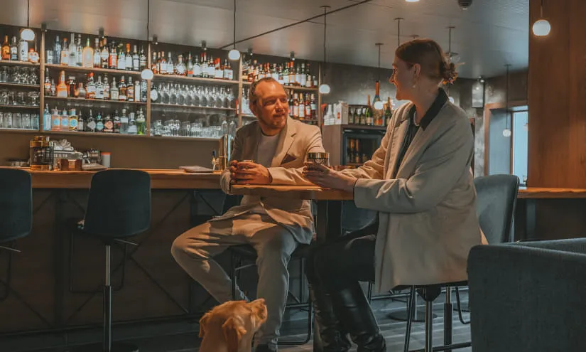 Zwei Personen unterhalten sich an einer Bar, ein Hund sitzt davor.